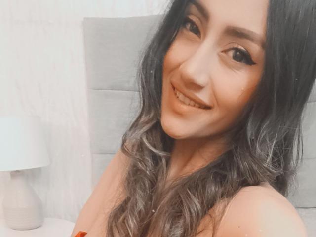 Helen Madisson, con cabello negro largo y ondulado, sonríe mostrando sus dientes, tiene ojos marrones y lleva una blusa roja, sentada contra un reposacabezas gris tapizado..