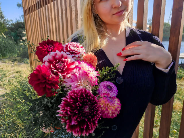 SallyVerda with blonde hair, red nails, wearing a dark cardigan, holding a bouquet of pink, red, and purple dahlias, standing in front of a wooden fence.