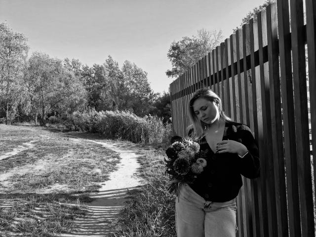 SallyVerda stands by a wooden fence, holding a bouquet of flowers, wearing a buttoned shirt and jeans, with a path and grassy area in the black-and-white shot.