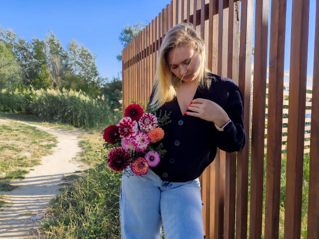 SallyVerda with blonde hair, wearing a black button-up and light blue jeans, holds a colorful bouquet of flowers, standing next to a wooden fence, looking down.