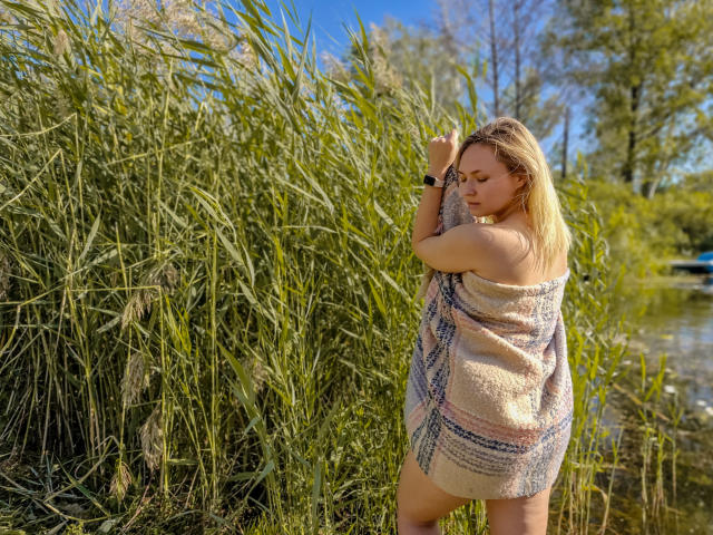 SallyVerda, with blonde hair, stands in tall grass, wearing a pink and blue patterned towel, covering her tits and ass. She's looking down, arm raised, by a clear pond.