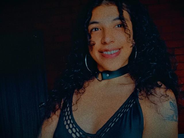 NicolWilliams with curly black hair, smiling, wearing a black mesh top and choker, showing small tits, hoop earrings, and a tattoo on her right arm. Dark room, dim light.