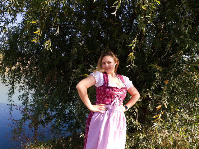 SallyVerda stands with hands on hips, wearing a pink and white Bavarian-style dress, her tits slightly visible, blonde hair, blue eyes, under a leafy green tree, by a water body.