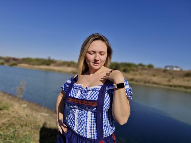 SallyVerda with blonde hair, blue and white checkered dress, small tits, wristwatch, standing by a river, eyes closed, smiling, sunlight, clear blue sky, grassy foreground.