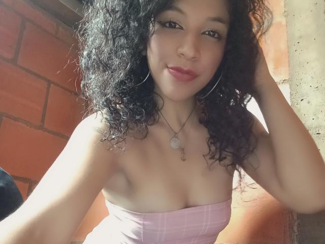 NicolWilliams with curly black hair, hoop earrings, pink strapless top, showing her tits, smiling at the camera, hand in hair, wearing a necklace with a circular pendant.
