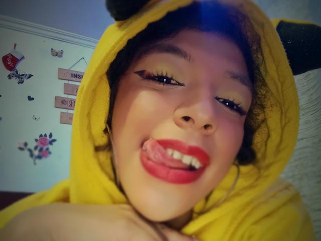 NicolWilliams with yellow Pikachu hoodie, tongue out, red lipstick, black eyeliner, yellow eyeshadow, smiling, holding her bare boob, curly dark hair, soft blue light, casual selfie.