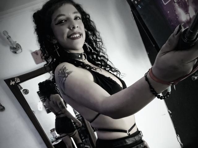 NicolWilliams with curly black hair, smiling, wearing a black choker, revealing tattoos, and a strappy black top, showing her tits, in a mirror selfie with a dark room.