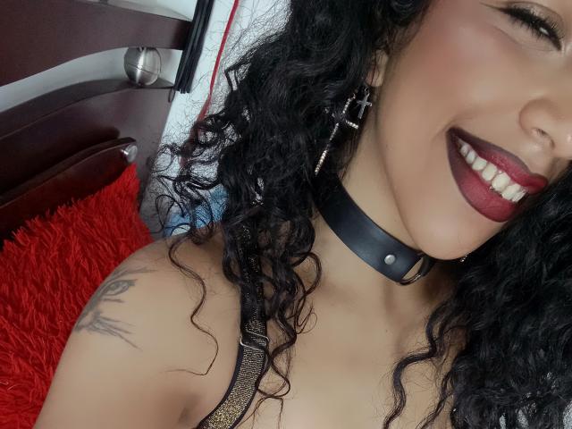 NicolWilliams with curly black hair, dark red lipstick, black choker, cross earrings, and tattoo on right shoulder, smiling at camera, showing her tits in a sparkly top.