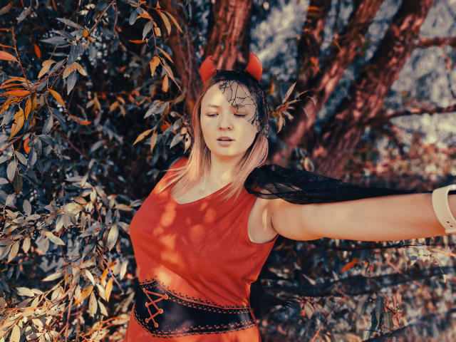 SallyVerda with blonde hair, red tank top, black lace headband with devil horns, black lace shawl, and corset, arms out, eyes closed, in sunlit forest.