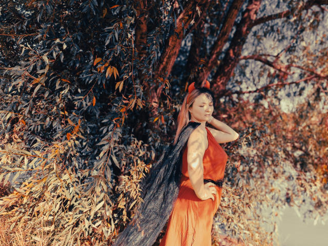 SallyVerda with orange dress, black fur cape, red fox ears, and hand on hip, standing against leafy tree, sunlight, and shadows, looking sideways, showing her ass.