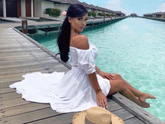 Hermosa y atractiva, con cabello negro largo, una pinza para el cabello rosa, un vestido blanco con un hombro descubierto, sentada en un muelle de madera, con las piernas en agua turquesa, un sombrero de paja en primer plano, mirando hacia atrás con una ligera sonrisa..