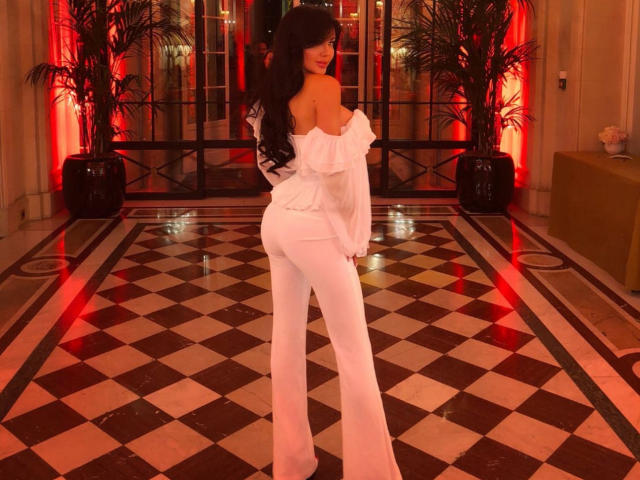 HotAndPretty with long black hair, off-shoulder white top, and white flared pants, showing her ass, standing on a checkered floor, red lighting in the background.