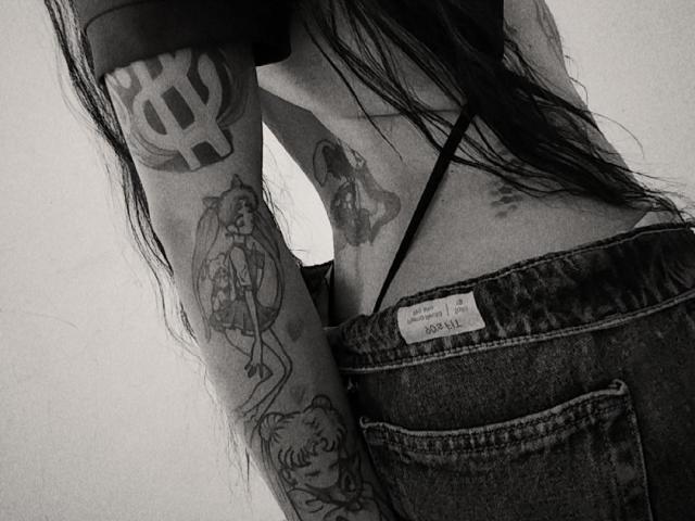 EvaMueller's back, showing tattoos on her arms and lower back, wearing denim with a black strap, long dark hair, black-and-white photo, visible tattoo on her right arm.