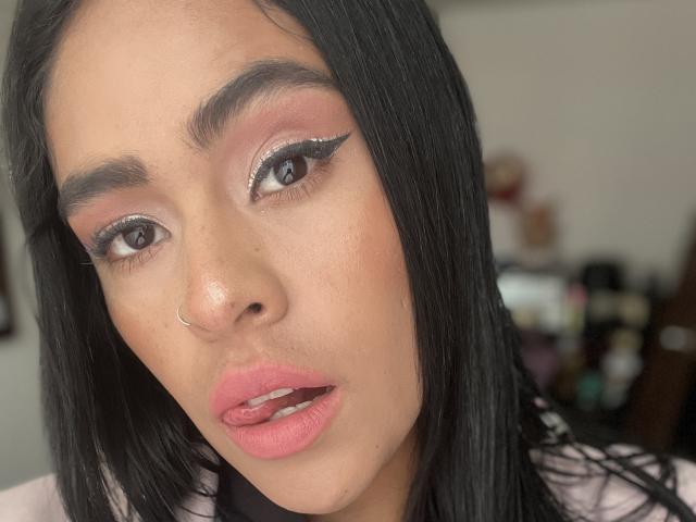 StellaBeckker has black hair, pink eyeshadow, black eyeliner with sparkles, brown eyes, pink lips, nose ring, and slightly parted pink lips, with a light brown skin tone.