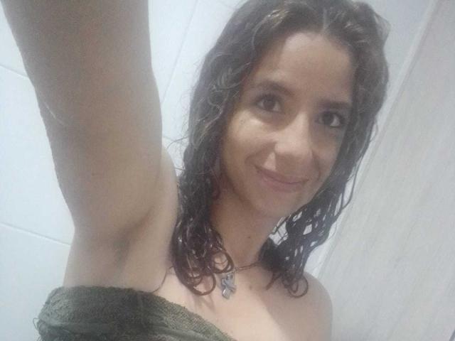 Sophie Teller, con el cabello castaño mojado, una toalla verde cubriendo sus pechos, sonriendo a la cámara, con un brazo levantado, axila visible, y llevando un collar con un dije..