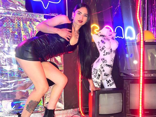 SarahMaxwell in a tight black leather mini-dress, long black hair, red lipstick, black ankle boots, tattoo on right thigh, standing next to a neon-lit mannequin with graffiti.