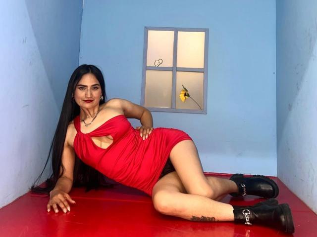 SarahMaxwell with long black hair, red dress, cutout, black boots, laying on red floor, blue walls, window, looking at camera, hand on floor, legs crossed, tattoo on thigh.