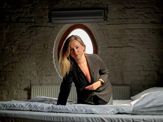 Holly-desire has blonde hair, a black dress, and is leaning on a bed with white sheets, looking at the camera with a slight smile, in a dimly lit room.