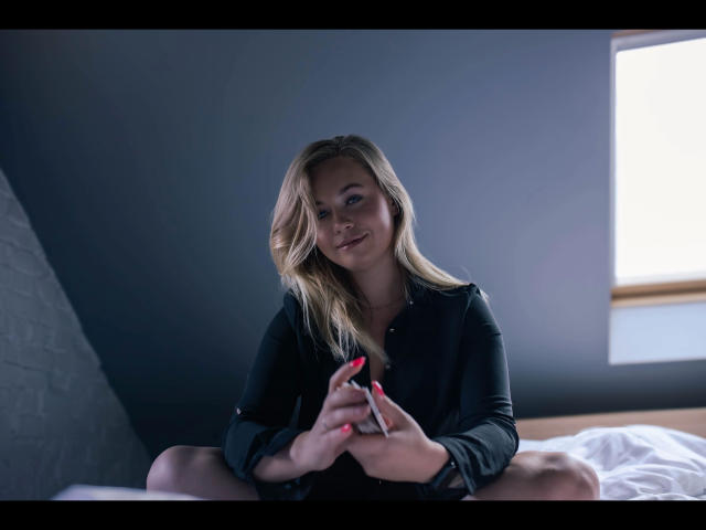 Holly-desire with blonde hair, smiling, sitting on bed, wearing black shirt, holding smartphone with red nails, legs spread, soft light from window.