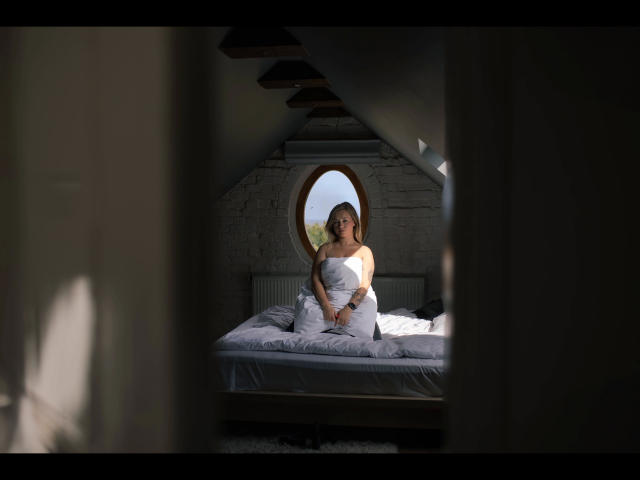 Holly-desire sits on a bed with white sheets, covering her tits with a blanket, looking through a circular window, in a dimly lit, slanted-roof room.