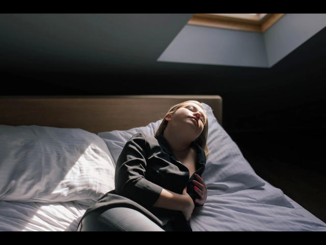 Holly-desire lies on a bed with white sheets, sunlit from a skylight, wearing a black shirt, eyes closed, head tilted back, hand on chest, looking relaxed and peaceful.