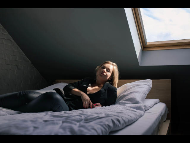 Holly-desire lying on a bed, wearing black leather pants and a black shirt, exposing her tits, with a skinned window above, looking relaxed and slightly aroused.
