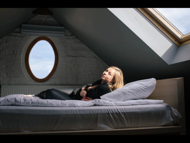 Holly-desire is lying on a bed with gray sheets, wearing black clothes, and looking up with a relaxed expression. She has blonde hair and a small ass. Skilled photo.