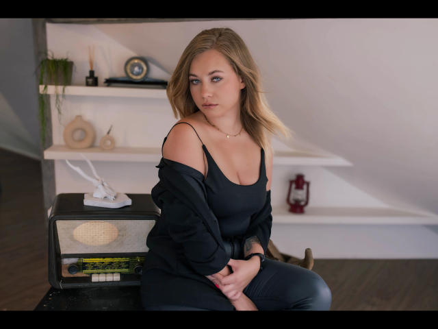 Holly-desire with blonde hair, blue eyes, and medium tits sits on a black radio, wearing a black off-shoulder top and black pants, in a softly lit room.