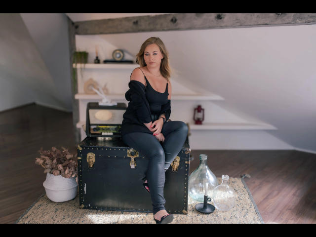 Holly-desire with bare shoulders, black off-shoulder top, and grey jeans, sitting on a black metal trunk, hands on knees, wooden floor, small glass bottles, and plant in front.