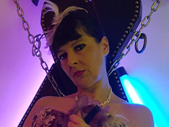 MaitresseElsa met kort zwart-wit haar, die paarse bloemen vasthoudt, een top zonder bandjes draagt en een ketting om haar nek heeft, zittend in een donkere leren stoel met metalen kettingen..