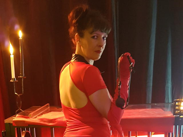 MaitresseElsa with red latex dress, back cutout, and matching gloves, holding a black strap, standing in front of a table with a candle, looking over her shoulder.