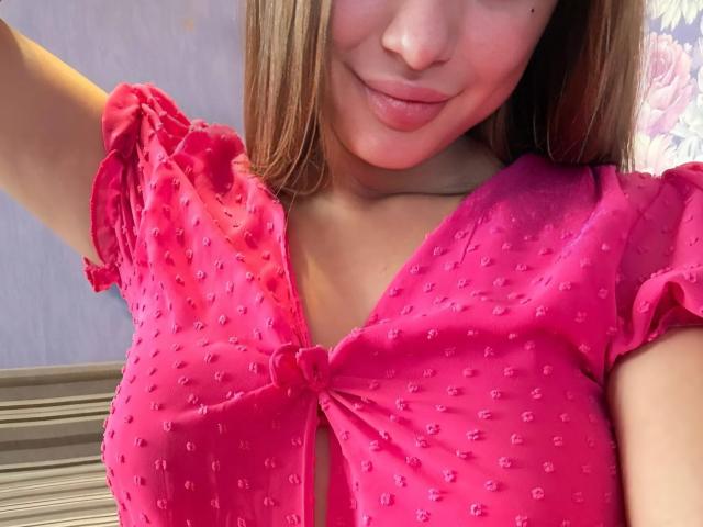 PeonyAmazing with pink, polka-dotted, sheer top, tied at the front, showing her small, cute tits, smiling with light brown hair, fair skin, and pink lips.