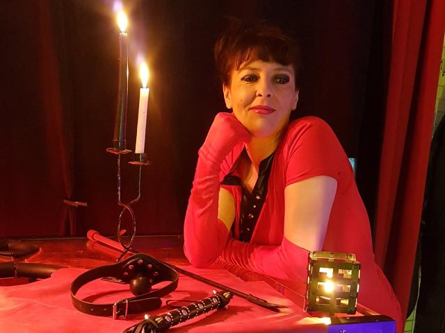 MaitresseElsa mit kurzen, braunen Haaren, einem roten Kleid und passenden Handschuhen, die an einem Tisch lehnt, auf dem eine Kerze brennt, mit einer schwarzen Maske und einem Lederriemen, wirkt sie verführerisch..