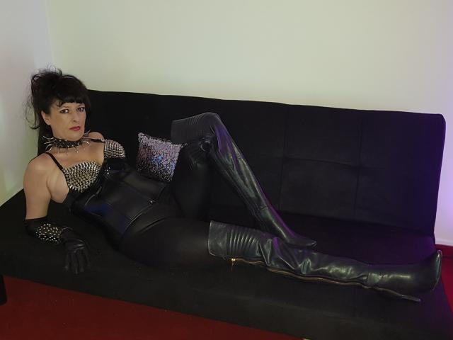 MaitresseElsa lounges on a black sofa in a leather bodysuit, thigh-high boots, and black gloves, showing her tits and ass, with a sparkly pillow beside her.