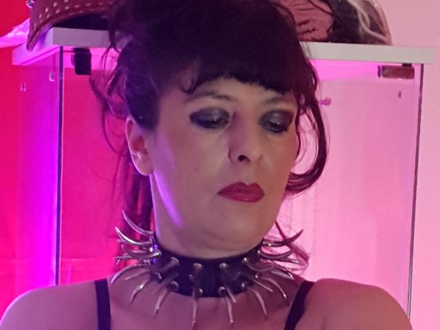 MaitresseElsa met donker haar, veel make-up, rode lippenstift, een zwarte choker met stekels en een zwarte jurk met bandjes, kijkt naar beneden met een serieuze uitdrukking, met roze en rode verlichting..