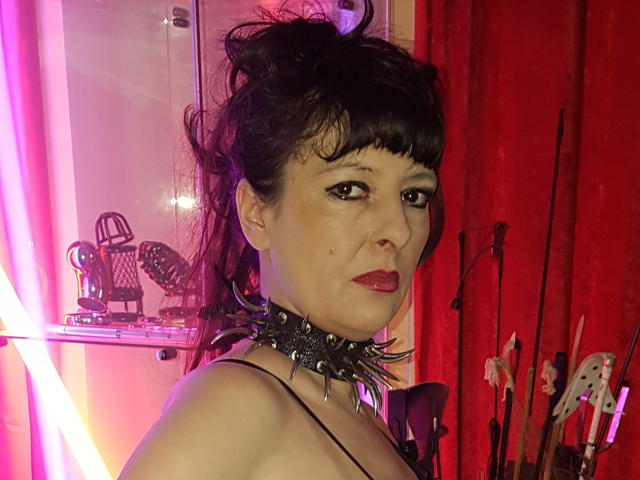 MaitresseElsa, con cabello oscuro, flequillo, lápiz labial rojo, collar con púas y hombros desnudos, mirando a la cámara, rodeada de luces neón rosa y rojas, con instrumentos musicales en el fondo..