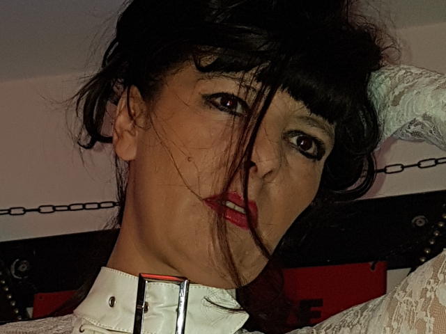 MaitresseElsa, com cabelo escuro, batom vermelho, camisa de renda branca e coleira de couro branco, olhando sedutivamente para a câmera, com uma corrente ao fundo. Seu rosto tem um leve sorriso..