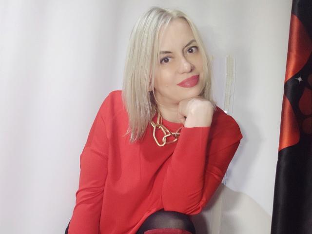 RebeccaDarling69 with blonde hair, red lipstick, and red top, wearing black tights, resting chin on hand, gold necklace, smiling, white background, partial American flag on right.