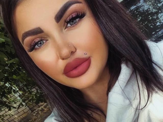 SophiaCaprice has dark brown hair, bold eyebrows, blue eyes, red lipstick, a nose ring, and a lip piercing. She's wearing a white jacket with a leafy background.