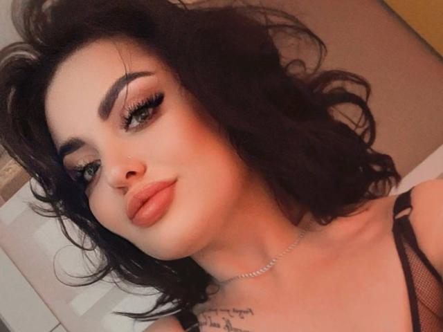 SophiaCaprice with dark wavy hair, full eyebrows, long eyelashes, and orange lipstick, wearing a black mesh top, showing a tattoo on her chest.