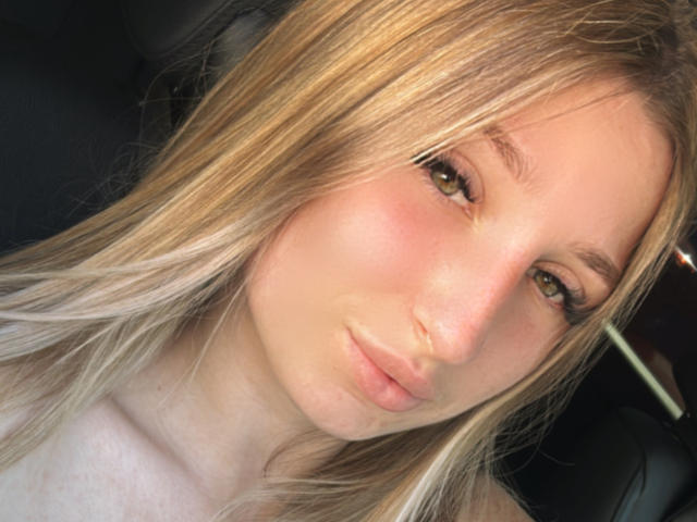 Le visage de SaraKeyt sur une photo, cheveux blonds, yeux verts, joues roses, lèvres légèrement entrouvertes de couleur rose, lumière douce, aspect naturel, maquillage discret, gros plan, siège de voiture en arrière-plan..