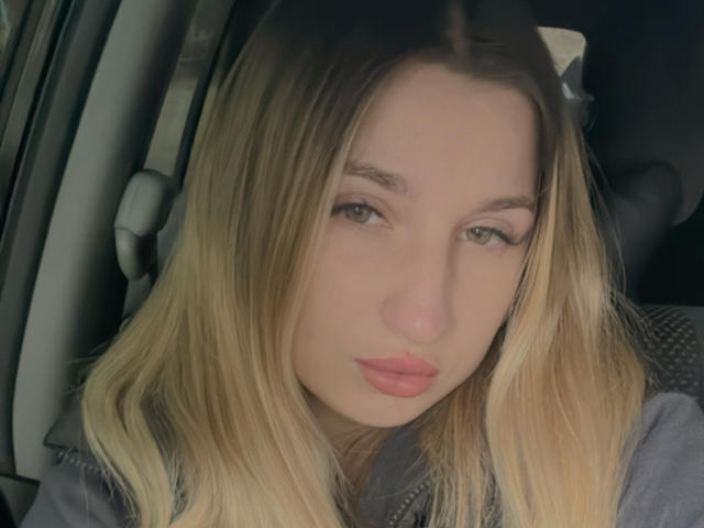 SaraKeyt, con i capelli biondi, le labbra rosa carnose e gli occhi verdi, è seduta in un'auto e guarda direttamente la fotocamera. Indossa una giacca grigia..