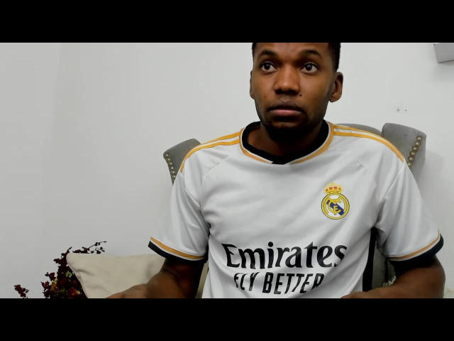 BryanOConner with dark skin, short black hair, wearing a white Real Madrid soccer shirt, looking surprised with a slight frown, hands resting on a grey chair.