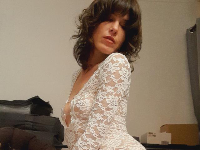Aiyana with wavy dark brown hair, wearing a white lace bodysuit, exposing her small tits and cleavage, standing in a dimly lit room, looking slightly to the side.
