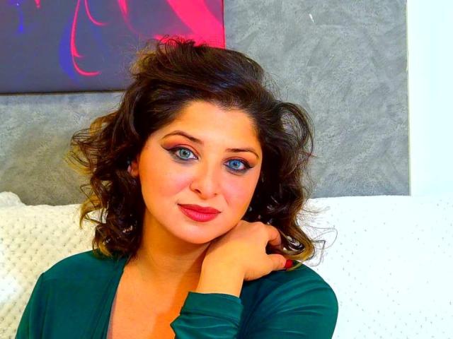 ScarletOra, con i capelli castano scuro ricci, gli occhi azzurri e il rossetto rosso, indossa una maglia verde, appoggia il mento sulla mano e guarda la fotocamera con un leggero sorriso..