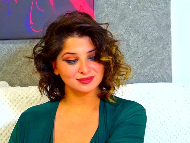 ScarletOra with curly brown hair, green dress, red lipstick, and blue eyeliner, smiling slightly, sitting on white textured pillow. Her tits are not visible.