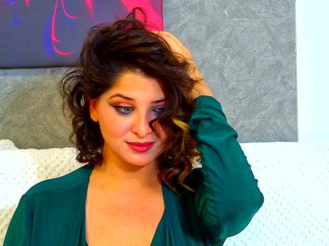 ScarletOra, con i capelli castano ricci, un vestito verde, rossetto rosso e una mano tra i capelli, guarda la fotocamera con un sorriso appena accennato, con il seno parzialmente visibile, in un primo piano..