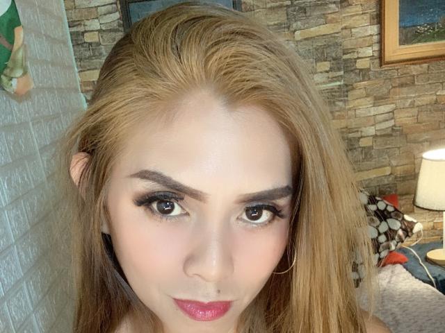 FingerMeForFun has blonde hair, brown eyes, black eyeliner, pink lipstick, and a slight smile. Her tits are partially visible. She's wearing gold hoop earrings.
