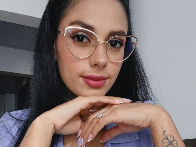AnaisHilton with long black hair, wearing pink lipstick and clear glasses, hands near face, showing a ring and tattoo, wearing a purple shirt. Her ass is not visible.