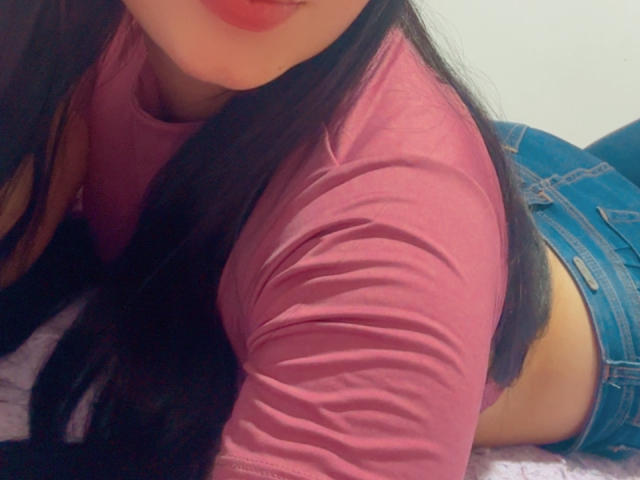 SarahGood lying on her stomach, pink shirt, blue jeans, long black hair, red lipstick, visible butt, soft bed, slightly blurred image.
